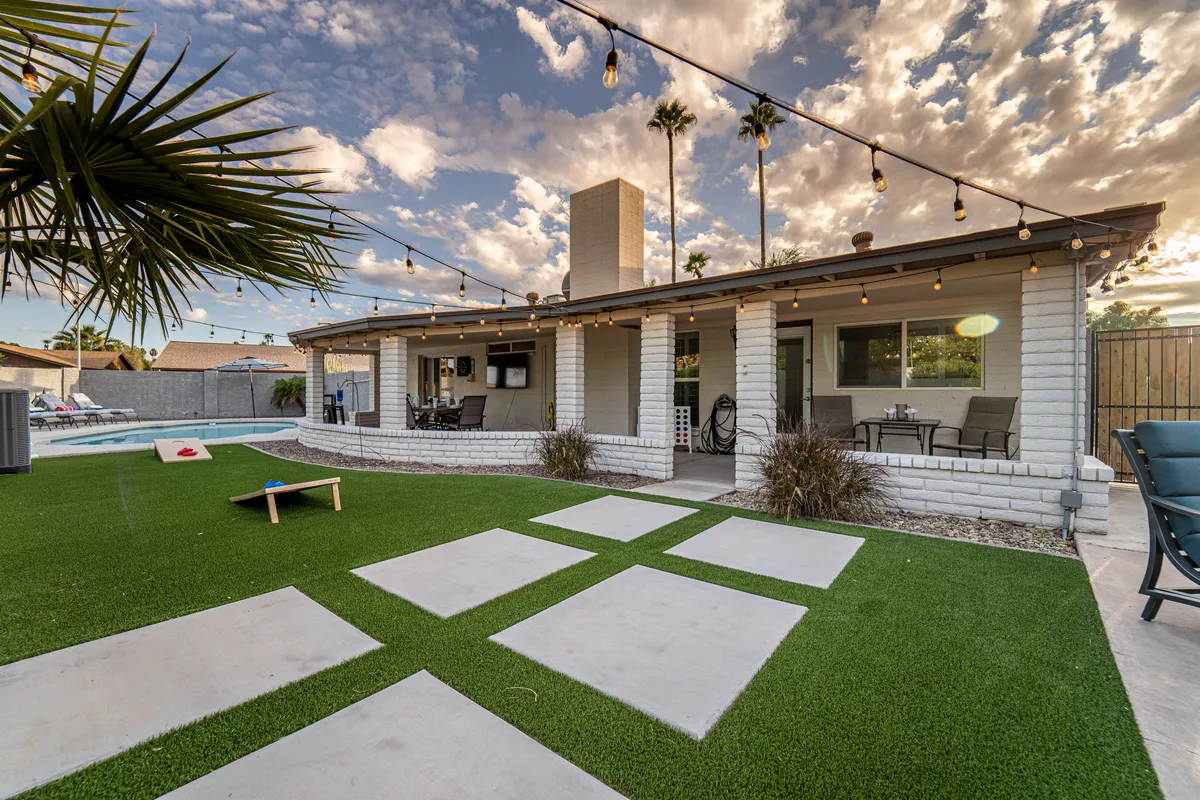 A vibrant backyard patio scene with a swimming pool, string lights overhead, and a welcoming outdoor seating area. The setting is festive yet serene, ideal for both daytime lounging and lively evening gatherings.