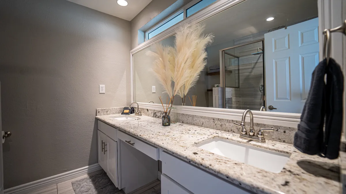 A spa-like bathroom featuring a granite countertop with dual sinks, a large mirror, pampas grass arrangement, and soft, tranquil lighting. The space feels serene and is ideal for self-care routines or unwinding after a long day.