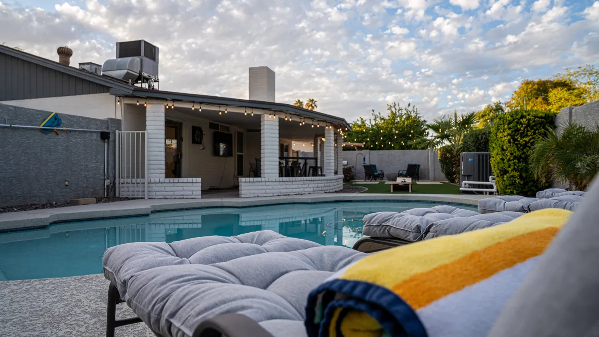 A sparkling swimming pool surrounded by lounge chairs and an outdoor seating area, all gently illuminated by string lights. The setting transitions beautifully from day to evening, with the sky painted in sunset hues. The overall mood is relaxed and resort-like, emphasizing spaces for swimming, sunbathing, and evening gatherings.