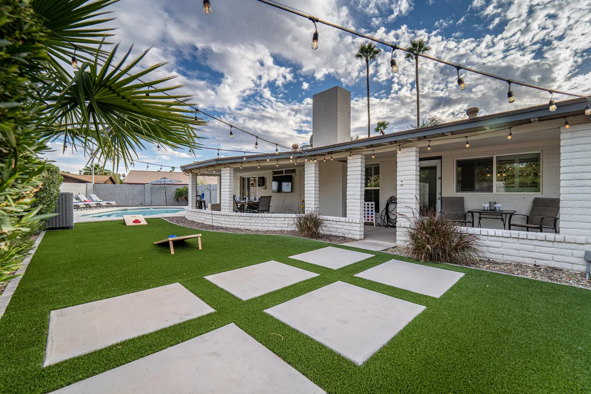 This image captures the heart of the backyard with a sparkling swimming pool surrounded by patio furniture, string lights overhead, landscaped grass, and a view of the house exterior. The scene conveys a relaxed, social atmosphere ideal for poolside lounging, dining alfresco, and enjoying yard games in the sunshine or under evening lights.