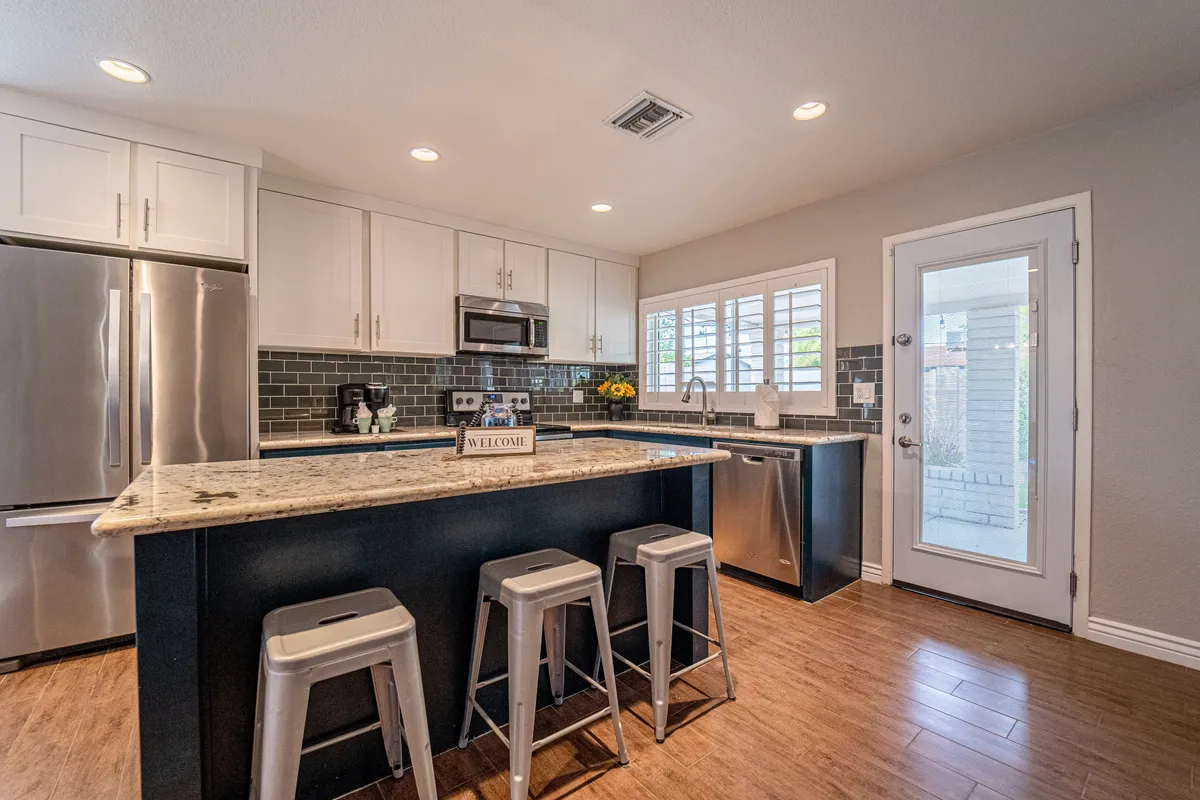 A bright, welcoming kitchen scene with granite countertops, stainless steel appliances, bar stools at the breakfast bar, and decorative flowers. Sunlight pours in, highlighting the inviting setup for cooking, serving, and socializing.