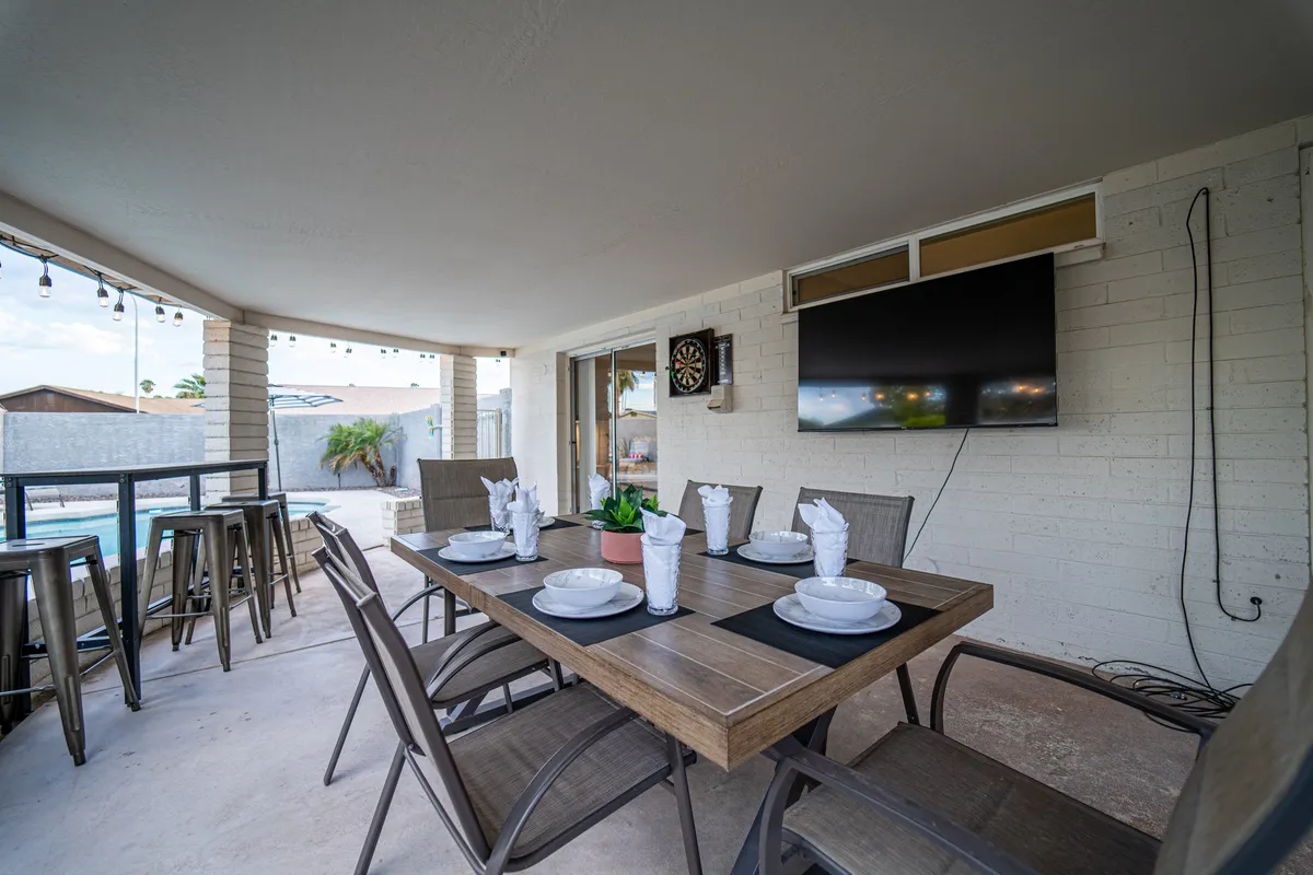 A vibrant outdoor dining and entertainment area with a table set for guests, cozy seating, a wall-mounted TV, dartboard, and direct views of the pool. The setup invites socializing, outdoor meals, and casual relaxation with poolside ambiance.