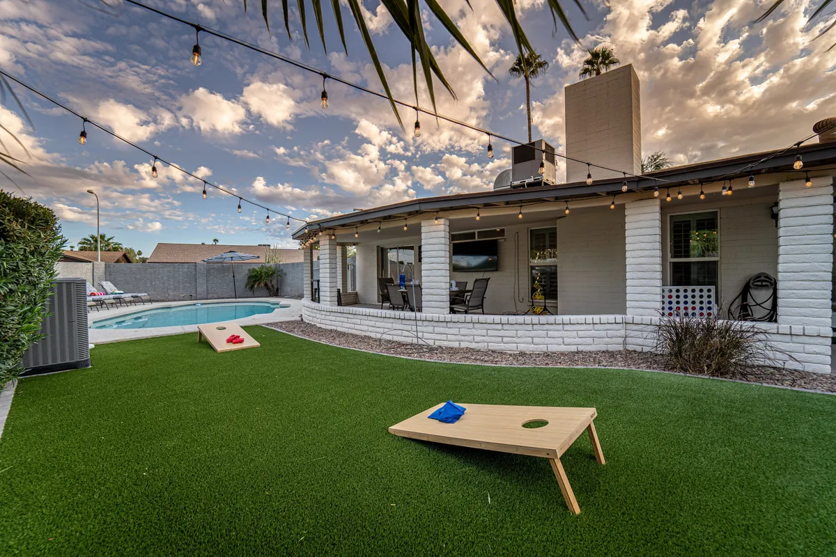 A backyard scene featuring a swimming pool, cornhole boards set up for a friendly game, string lights overhead, and a cozy outdoor seating area. The area is set for both relaxation and playful activities, ideal for gatherings and competitions at dusk.