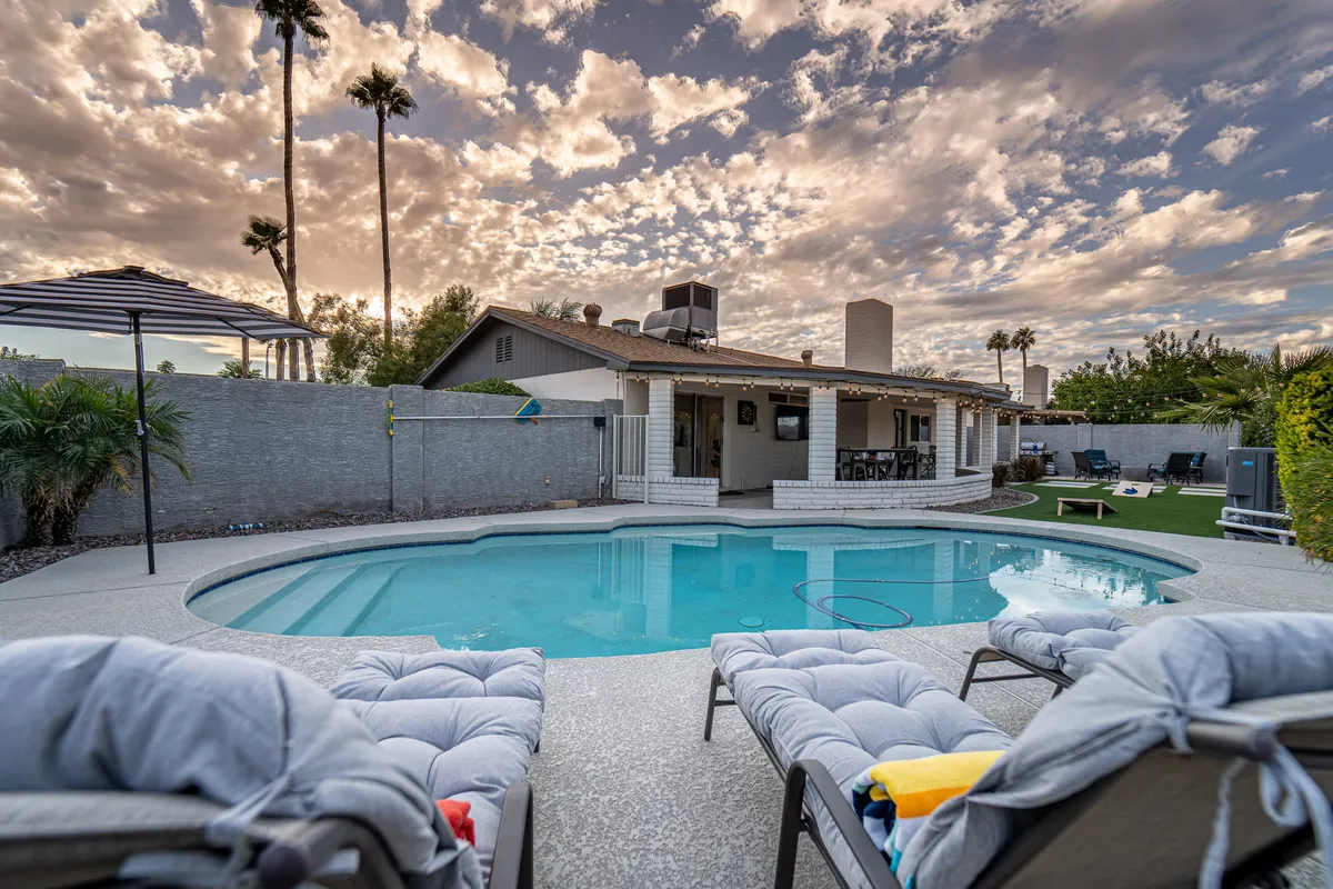 A tranquil backyard scene featuring a swimming pool surrounded by lounge chairs and palm trees, with the house exterior visible in the background. The area is bathed in soft light, suggesting a relaxing and leisurely atmosphere ideal for swimming, sunbathing, or social gatherings.