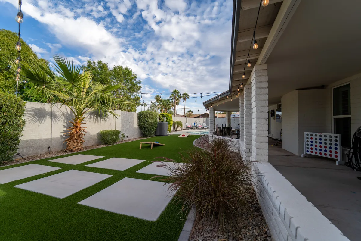 The backyard is showcased with a green lawn, graceful palm trees, glowing string lights, a patio space, and room for outdoor games. The atmosphere is lively and social, suggesting the ideal setting for gatherings after a day spent exploring nearby attractions.