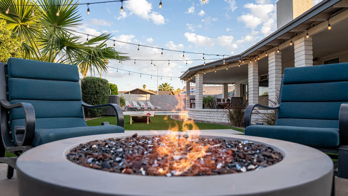 An outdoor lounge area featuring a glowing fire pit, surrounded by comfortable lounge chairs and illuminated with delicate string lights. Palm trees and an open sky create a cozy, intimate setting for evening gatherings or quiet reflection.