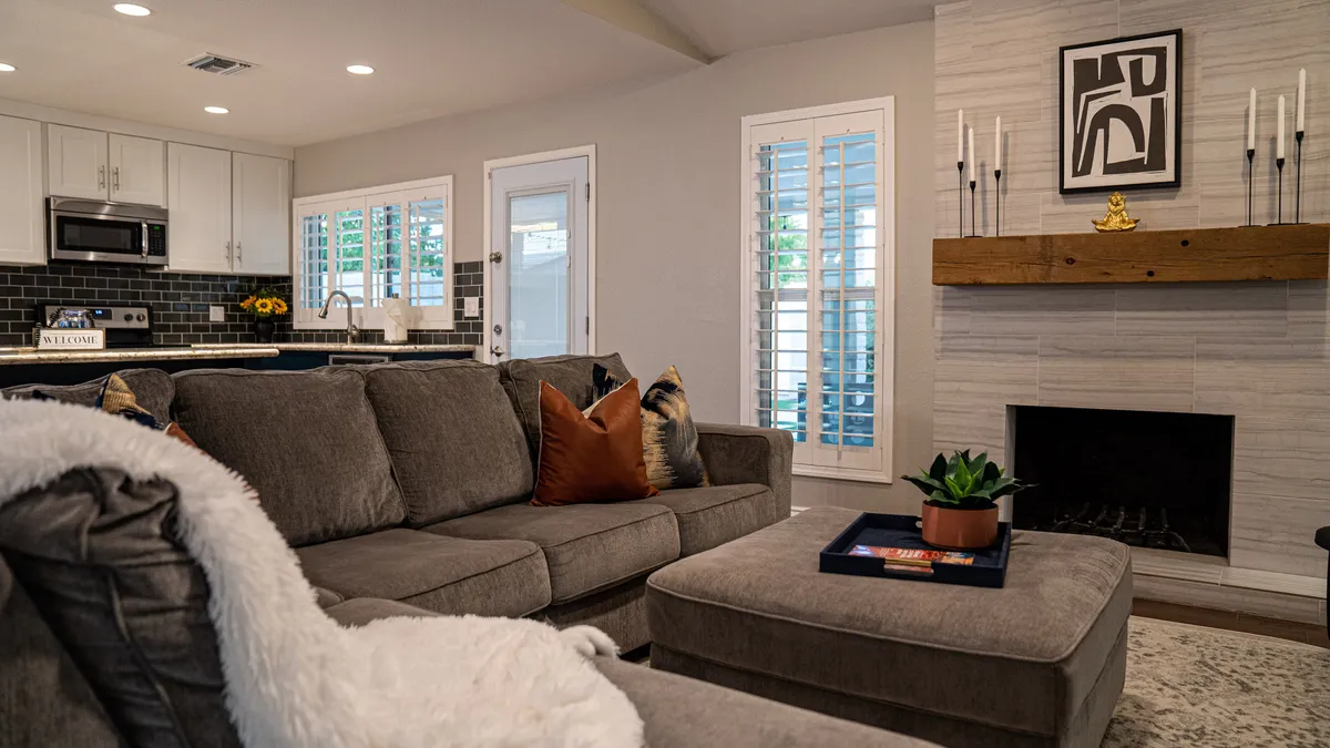 A spacious, open living area with a large, plush gray sectional sofa as the centerpiece, accented by stylish pillows. In the background, a modern kitchen is visible, and a wooden mantelpiece with decorative items adds warmth and subtle sophistication.