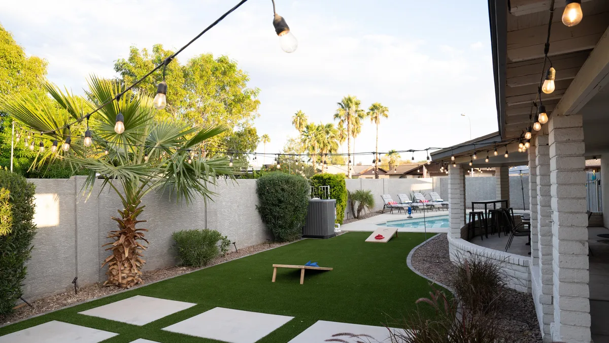 The image presents a vibrant backyard scene featuring a swimming pool, string lights crisscrossing the sky, a lush lawn, palm trees, and lawn games set up nearby. The setting invites both daytime play and evening gatherings, with the pool at the center and ample space for groups to relax.