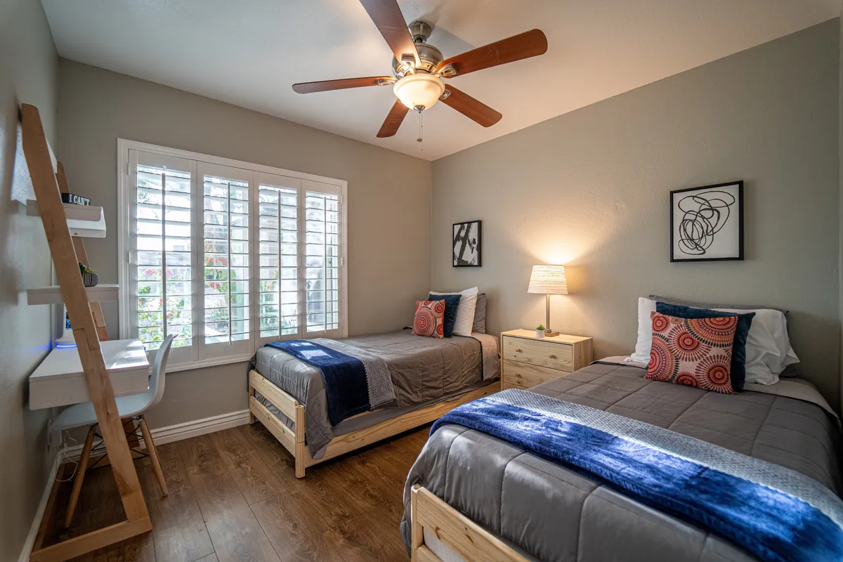A guest bedroom with two twin beds (convertible to a king), a desk and chair, a window with shutters, and a lamp. The room is cozy, versatile, and equipped for both relaxation and practical needs such as reading or studying.
