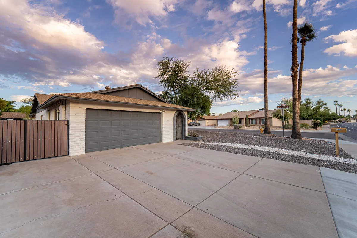 A view of the garage door framed by an entrance archway and shaded by palm trees, suggesting easy access and ample space for parking. The scene is inviting and highlights the exterior functionality of the property.